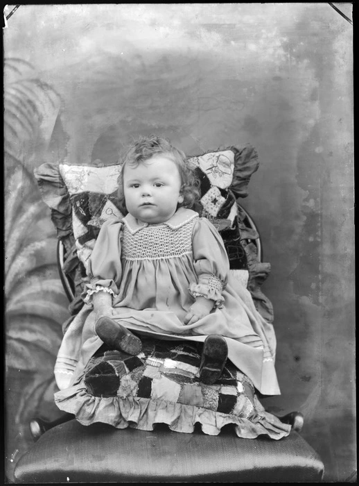 Studio portrait of unidentified infant wearing a smocked dress and sitting on and against patchwork cushions, probably Christchurch district