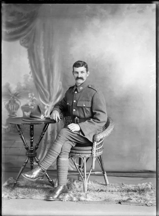 Studio portrait of unidentified soldier with moustache in uniform wearing putties, sitting in cane chair at table with hat, Christchurch