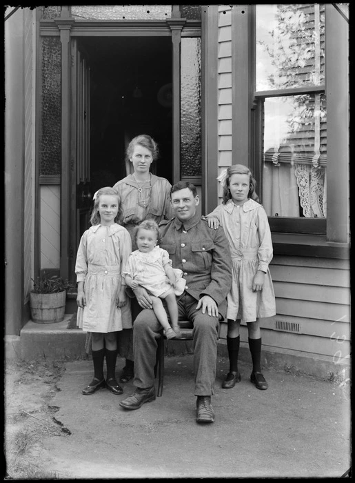 Family unidentified portrait in front of wooden house, father in soldier's uniform sitting with toddler daughter, mother with necklace standing with older two daughters, probably Christchurch region