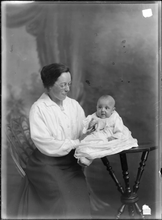 Studio family portrait of an unidentified woman with glasses and brooch, sitting holding a baby in christening gown on a table, Christchurch