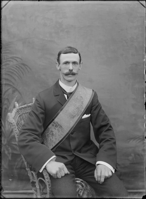 Studio portrait of an unidentified member of the Independent Order of Oddfellows Manchester Unity, dressed in a suit and tie and wearing a sash, sitting on a cane chair, possibly Christchurch district