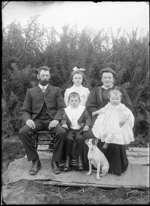 Outdoors unidentified family portrait in front of gorse hedge, parents wearing glasses sitting, father with beard, mother with baby on knee and young son with dog between, older daughter behind, probably Christchurch region