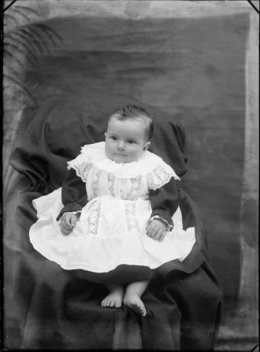 Studio portrait of unidentified baby wearing a lace dress seated on a covered chair, probably Christchurch district