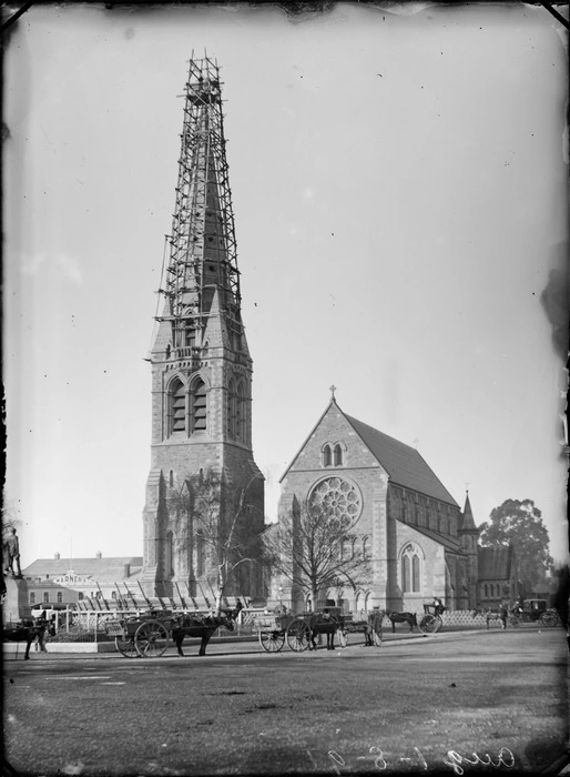 View of the Cathedral Church steeple under repair from prior earthquake damage, with John Robert Godley statue and horse drawn wagons and coaches in foreground, with Warner's Hotel beyond, Cathedral Square, Christchurch
