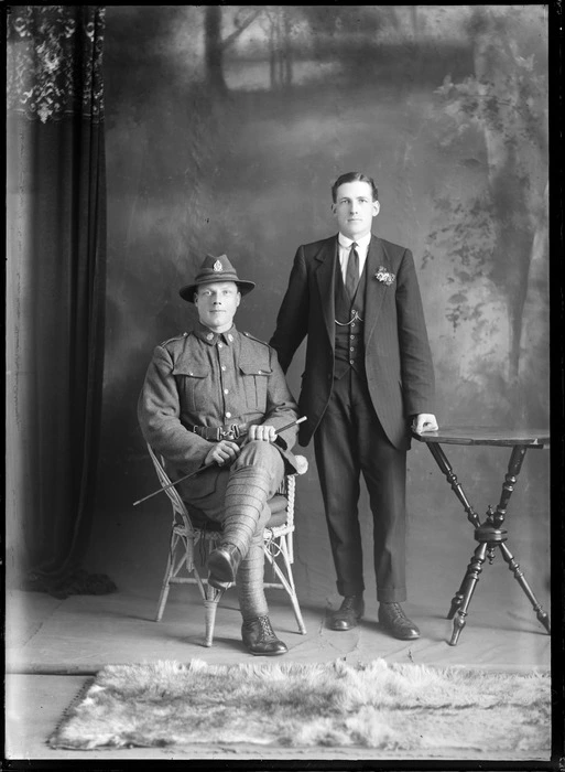 Studio portrait of two unidentified men, soldier seated in uniform and the other standing with hand leaning on table, probably Christchurch district