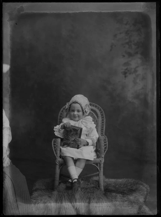 Studio unidentified family portrait of a young girl in a lace collar and sleeve corduroy dress and bonnet with bow, sitting in a cane chair with a cat book, mother just in view, Christchurch