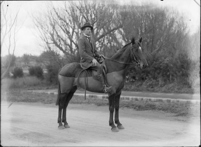 Unidentified man in riding boots, stirrups and hat, astride a chaser clip groomed show jumping horse, with whip hanging from saddle, on a dirt road in front of trees, probably Christchurch region