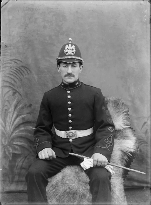 Outdoors in front of false backdrop, portrait of an unidentified soldier in dress uniform, with moustache, collar badges, sleeve insignia, wearing a domed helmet with large lion and unicorn badge, probably Christchurch region