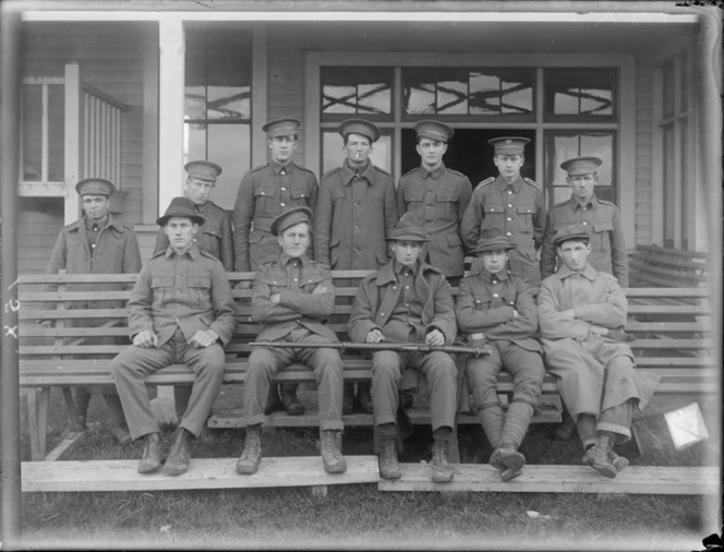 A group of unidentified soldiers in front of the [barracks?], five sitting a wooden bench, with a rifle on the laps of two soldiers, at casual camp, Hastings