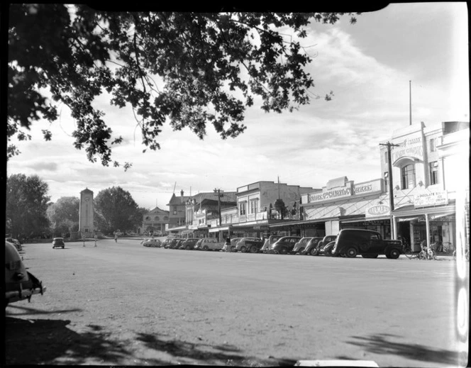 Street scene, Cambridge, Waikato region, with cars, clock tower and shops, including premises of drapers and mercers Geo Calvert and Co
