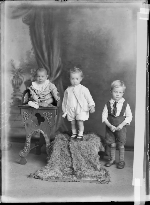 Studio portrait of unidentified children, young boy with large collar, tie and braces, standing next to his toddler sister and a baby wearing a bib on a high chair, Christchurch