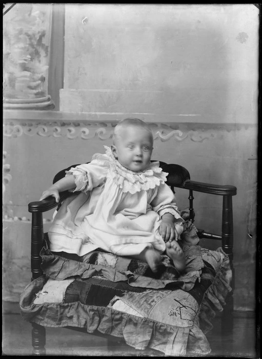 Portrait of an unidentified infant, sitting on patchwork cushion, wearing a gown with a ruffled collar, possibly Christchurch district