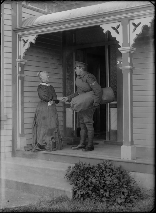 Outdoors on a veranda of a wooden house, a farewell between an unidentified elderly mother with her older World War I Corporal Soldier son carrying his coat and kit bag, probably Christchurch region