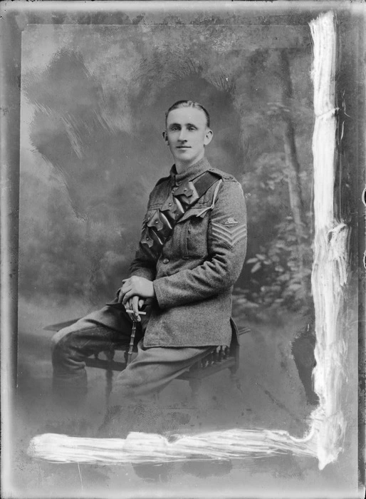 Studio portrait of unidentified World War I soldier with bandolier ammunition belt, artillery sleeve badge and white shoulder pocket cord, sitting with cigarette and swagger stick in hand, location unknown