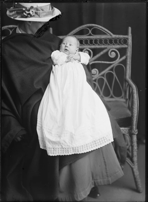 Studio portrait of an unidentified baby in a christening gown being held by their mother sitting with a blanket and straw hat on a cane couch, Christchurch
