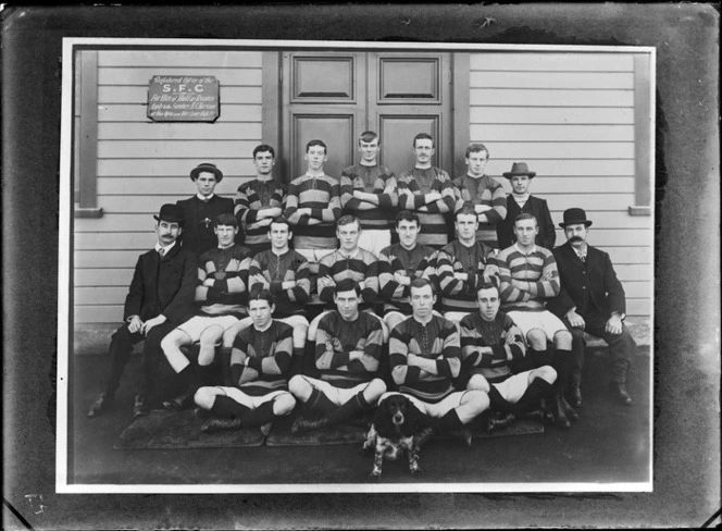 Unidentified rugby football team portrait in front of wooden building with 'SFC, H C Harrison' sign, includes fifteen players in uniform with four men in suits and a Cocker Spaniel dog, probably Christchurch region