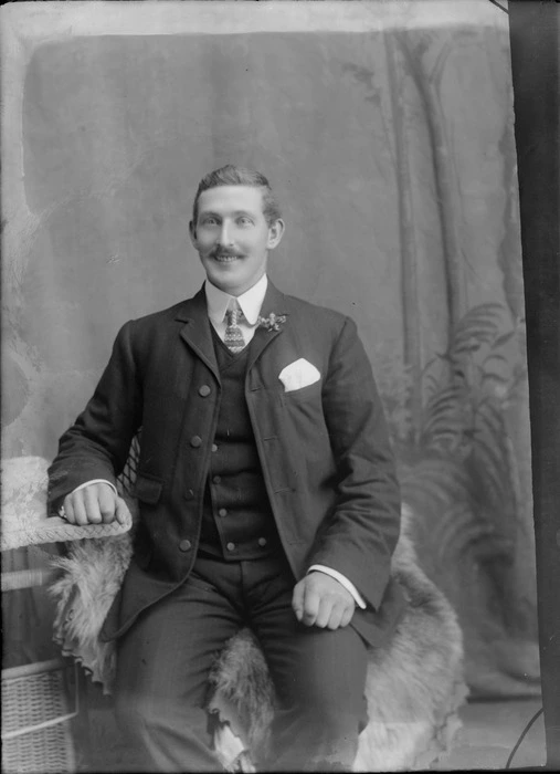 Studio portrait of an unidentified man with moustache, double round shirt collar and patterned tie, and three piece suit with lapel flower, sitting at a cane table, Christchurch