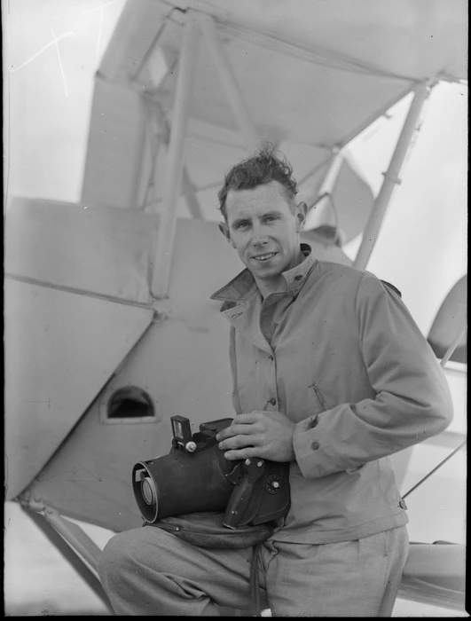 Portrait of Clyde (Snow) Stewart, Production Manager for Whites Aviation, holding a K 20 aerial camera in front of a Tiger Moth biplane