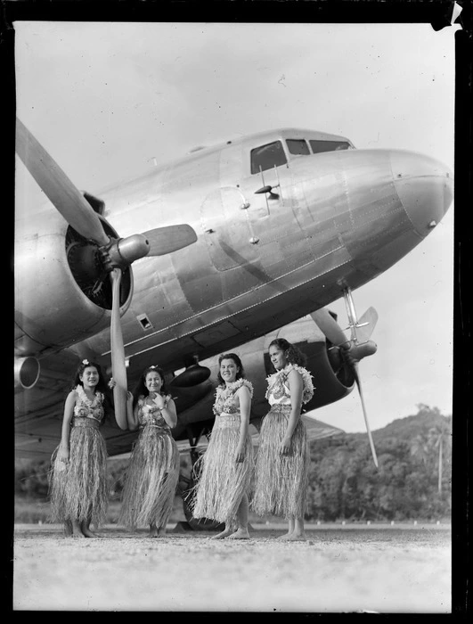 Four unidentified local girls wearing hula skirts in front of a C47 transport aircraft, Rarotonga airfield, Cook Islands