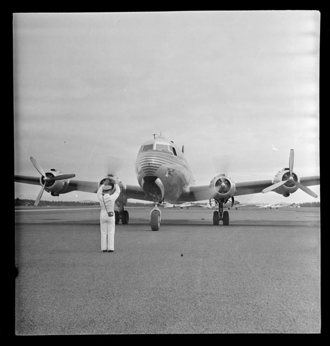 View of Pan American Airways Clipper Class Cathay DC4 passenger plane Good Will Flight taxiing on Whenuapai Airfield runway with unidentified ground personnel in front, Auckland