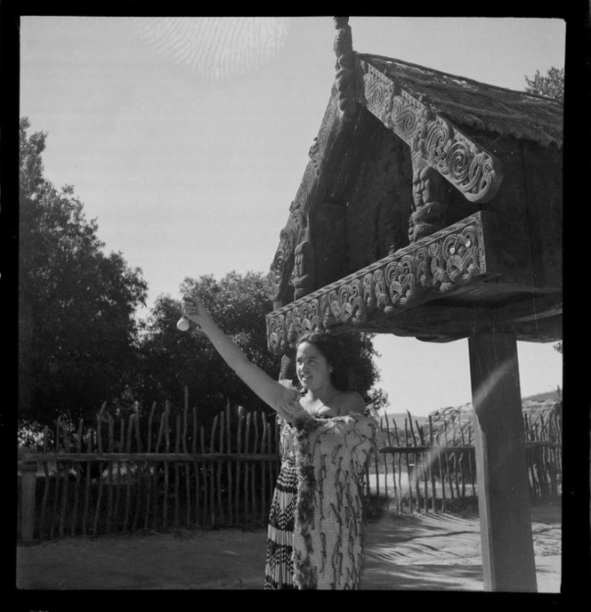 Unidentified Maori girl wearing piupiu and cloak, with poi standing under carved Pataka, Whakarewarewa, Rotorua