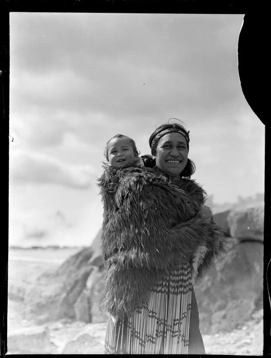 Unidentified Maori mother with a baby on her back wrapped in animal skin, woman is wearing a piupiu skirt