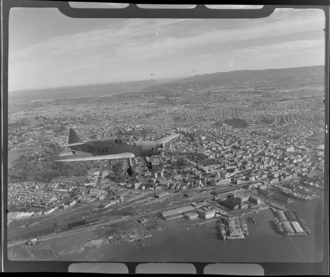 Auckland Aero Club's DH94 Moth Minor KZ-AJX plane in flight with Auckland City and waterfront in view