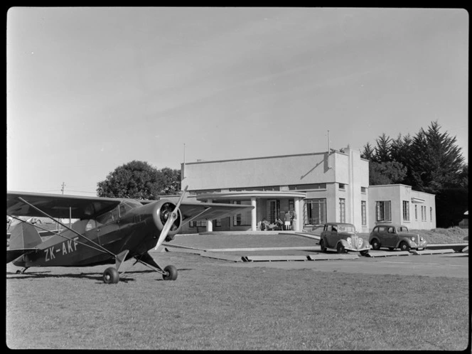 View of the clubrooms at Auckland Aero Club, Mangere, Auckland, showing Rearwin Sportster aeroplane ZK-AKF and two motorcars parked outside