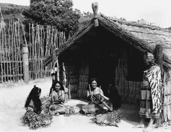 Maori women, in semi traditional costume, playing a stick game alongside a meeting house