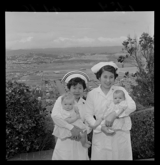 Portrait of two unidentified Far East Nurses holding babies at Karitane Hospital with Lyall Bay beyond, Melrose, Wellington City