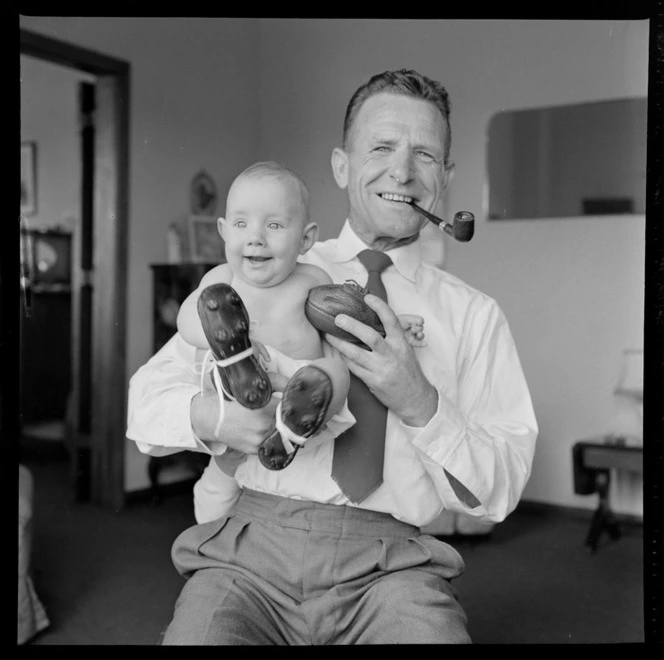 Neill McGregor holding miniature rugby ball and his baby grandson Neill Thomas McGregor wearing small sized rugby boots