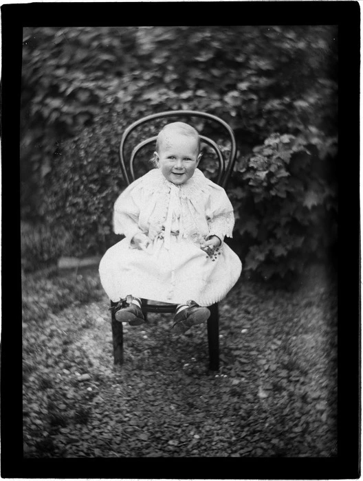 Edgar Williams as a baby sitting on a chair holding flowers in the garden of their home 'View Bank', Maitland Street, Dunedin