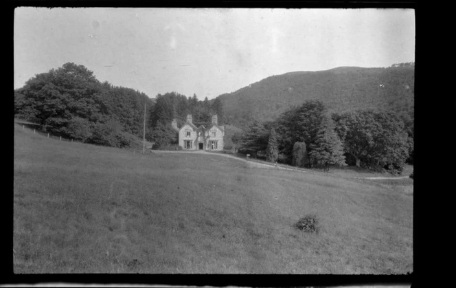 View over a field to a large two storey stone house with flag pole and road in front of a forest covered hill, Wales