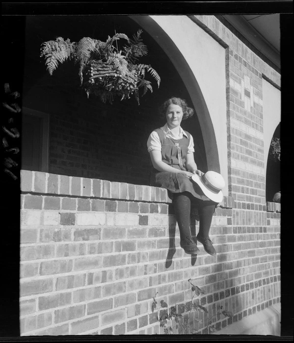 An unidentified schoolgirl sitting in an arched opening in a brick wall, Westport Technical High School, West Coast Region