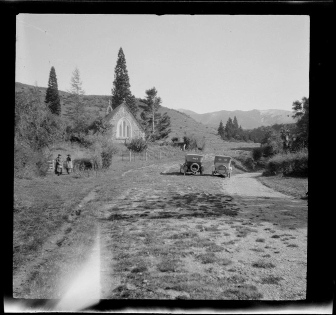 Stone chapel surrounded by trees, with two unidentified women standing beside church gate in the middle distance, and two motorcars parked next to road, Church of the Holy Innocents, Mount Peel Station, Canterbury Region