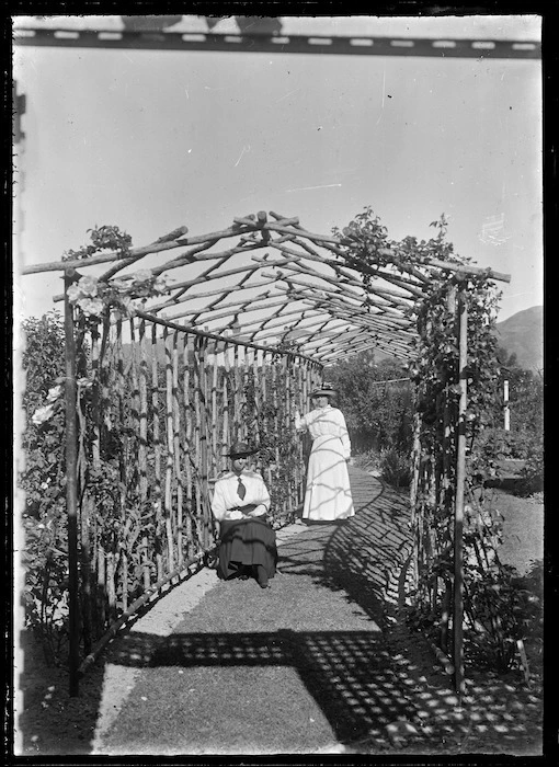 Two unidentified women in a latticed arbour with climbing roses.