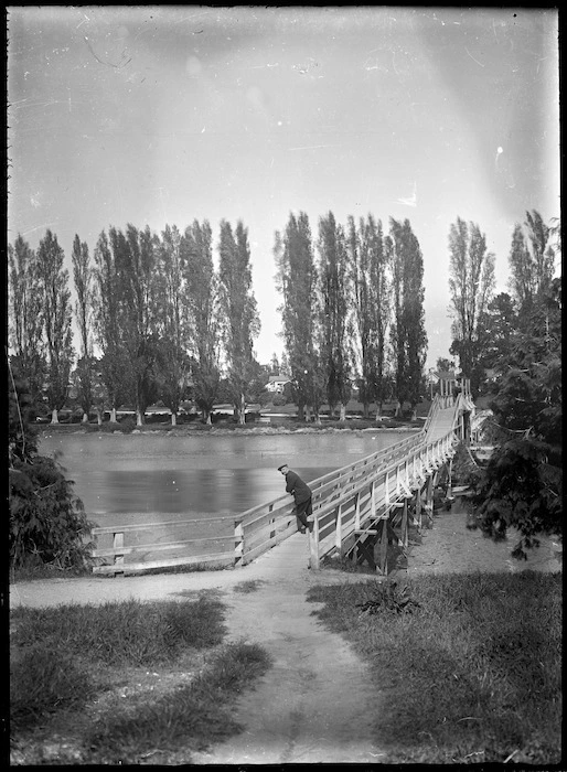 Albert Percy Godber standing on a footbridge over the Taruheru River, Gisborne, with a row of poplars on the far side.