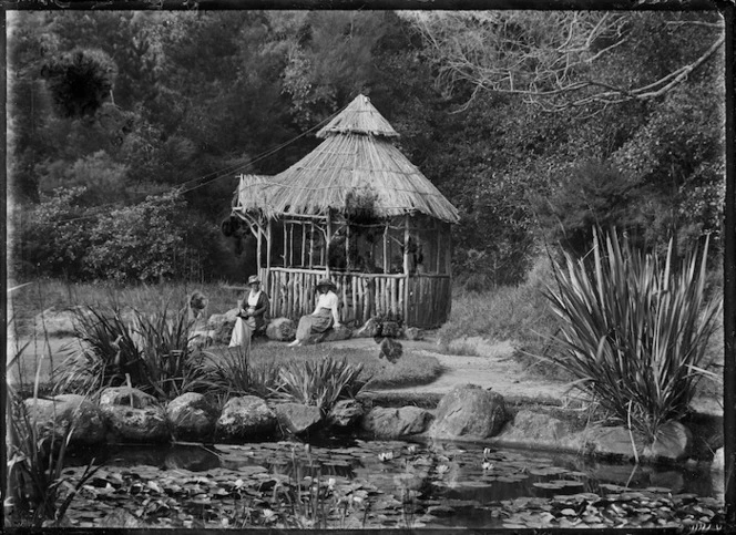 Two women seated on rocks in front of a thatched summer-house containing a drinking fountain at the Domain, Te Aroha, 1921