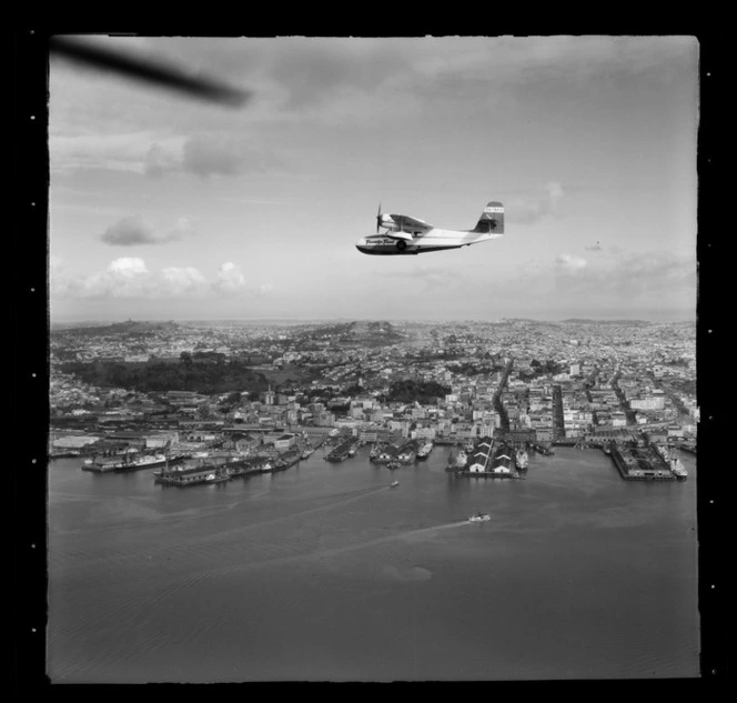 Tourist Air Travel, Grumman Widgeon aircraft in flight above Auckland Wharves and central city