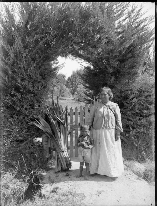 Unidentified Maori woman and child with a bundle of harakeke, outside a gate