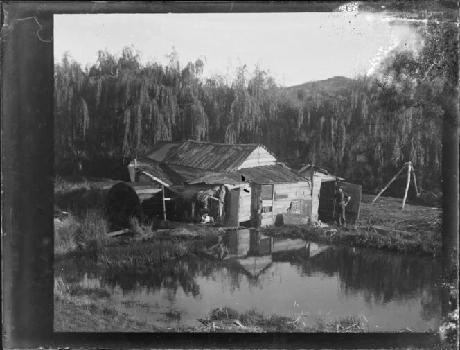 Two men and a dog, outside a wooden building next to a pond, location unidentified