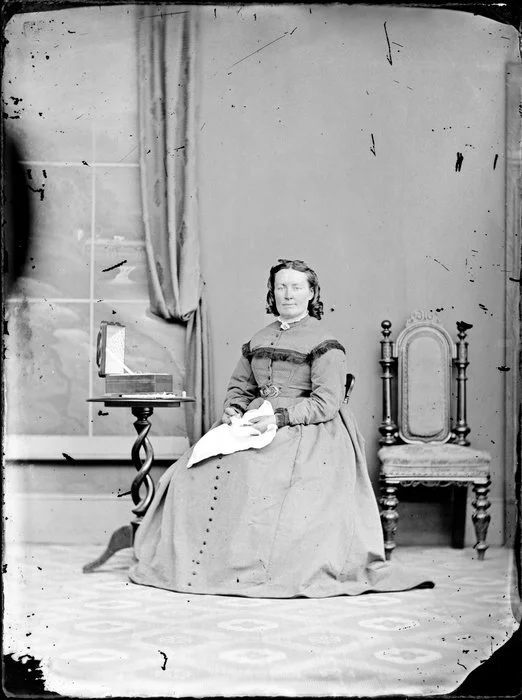 Unidentified woman seated, wearing crinoline dress with belt around the waist, tassels top part of dress and around the cuffs