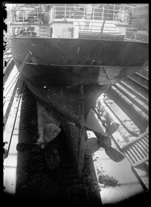 View of the stern of a ship showing the propellers with men working at the bottom of an unidentified dry dock