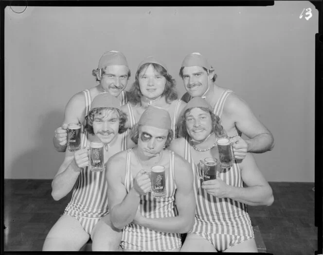 People (one with a fake black eye) in surf lifesaving costumes, drinking Lion beer in studio