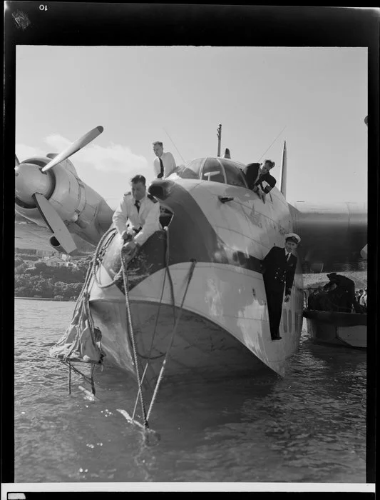 Tasman Empire Airways Limited crew watch on as the Short S.45 Solent flying boat, R.M.A Araragi (ZK-AMM), is moored by a man leaning out of the nose of the aircraft, Evans Bay, Wellington