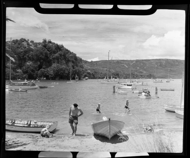 Unidentified child playing in the sand with a man watching while others swimm and boat, Whitianga harbour