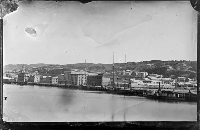 View of buildings on Customhouse Quay, Lambton, Wellington, including warehouses of W & G Turnbull and Company, and the Chief Post Office, with ships docked at the quay, and houses on the Terrace in background
