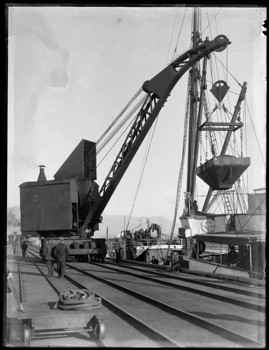 Wharf area, Westport, with crane on rails lifting coal hopper, ship and Westport Harbour beyond