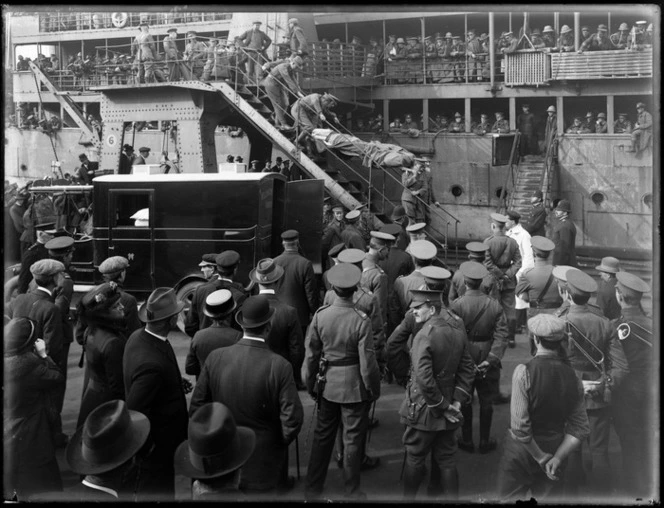 Invalided World War I soldiers arriving home on troop ship, one being carried on stretcher down some stairs to a waiting Wellington Ambulance, senior officers and army band and people waiting on wharf looking on
