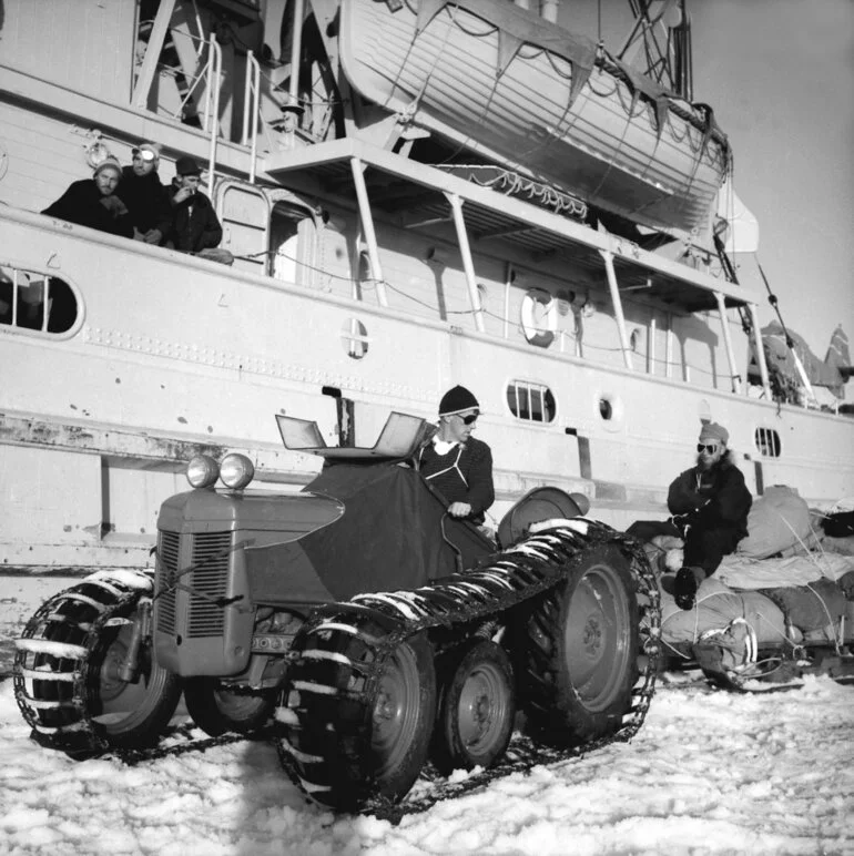 Image: Sir Edmund Hillary drives a tractor and stores away from the side of HMNZS Endeavour while IGY Scientific Leader Trevor Hatherton rides the sled